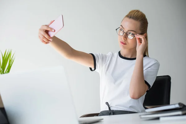 Bela jovem mulher em óculos tomando selfie enquanto sentado no local de trabalho — Fotografia de Stock