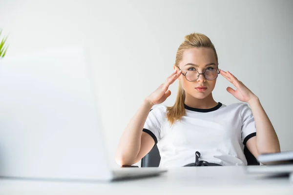 Beautiful young blonde woman in eyeglasses looking at camera while sitting at workplace — Stock Photo