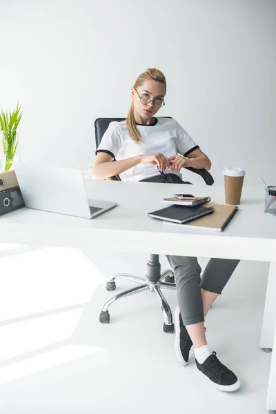 Hermosa joven mujer de negocios en gafas mirando a la cámara mientras está sentado en el lugar de trabajo - foto de stock