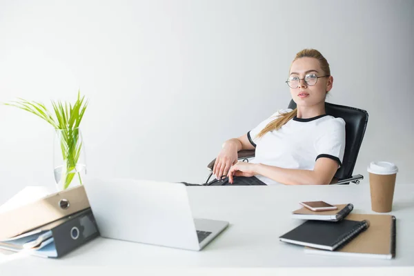 Beautiful young blonde businesswoman in eyeglasses looking at camera while sitting at workplace — Stock Photo
