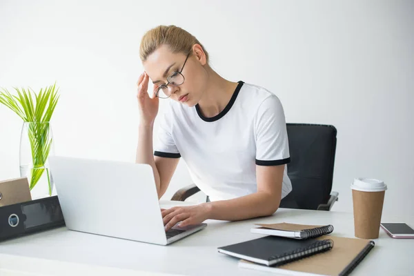 Tired young businesswoman in eyeglasses using laptop in office — Stock Photo