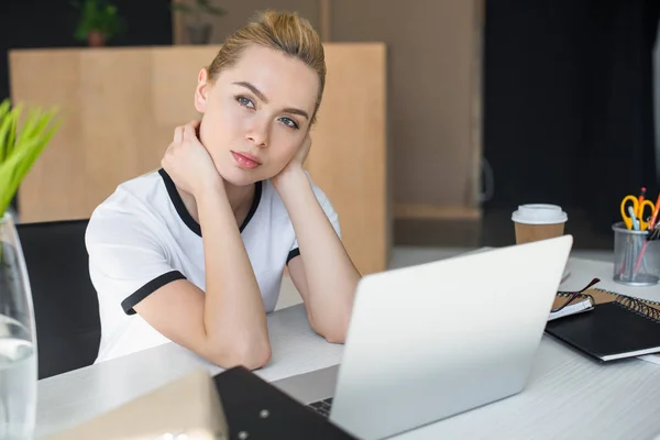 Задумчивая молодая деловая женщина смотрит в сторону, сидя за столом с ноутбуком в офисе — стоковое фото