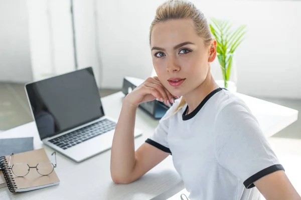 Belle jeune femme regardant la caméra tout en travaillant avec un ordinateur portable au bureau — Photo de stock