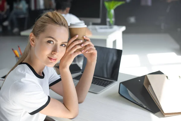 Hermosa joven empresaria sosteniendo taza de café desechable y mirando a la cámara en la oficina - foto de stock