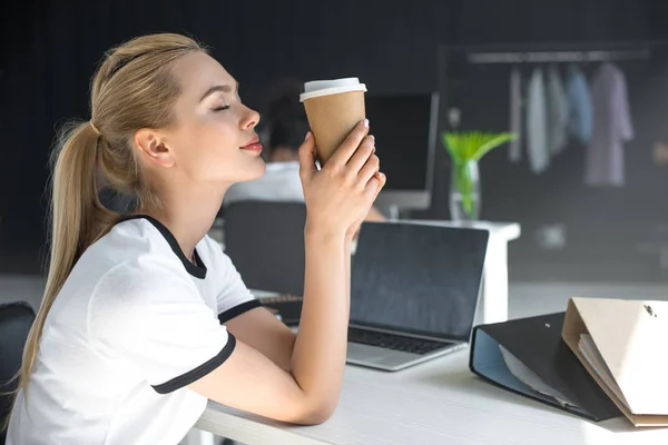 Боковой вид расслабленной молодой женщины с закрытыми глазами, держащей бумажную чашку в офисе — стоковое фото