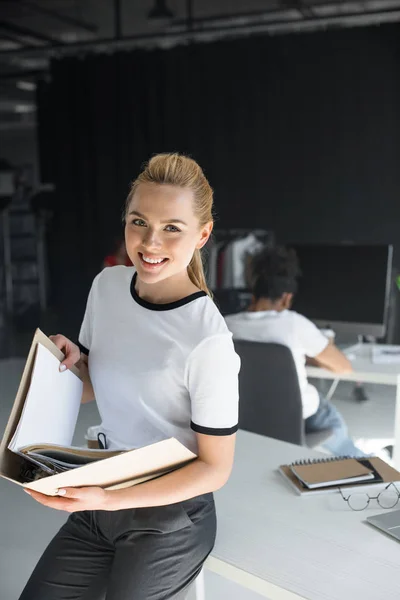 Attractive young businesswoman holding folder with documents and smiling at camera in office — Stock Photo