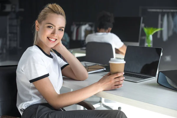 Belle jeune femme tenant tasse en papier et souriant à la caméra dans le bureau — Photo de stock