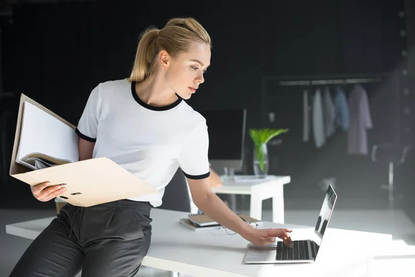 Hermosa joven que sostiene la carpeta y el uso de la computadora portátil en la oficina - foto de stock