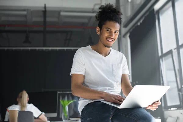 Junger afrikanisch-amerikanischer Mann benutzt Laptop und lächelt im Büro in die Kamera — Stockfoto