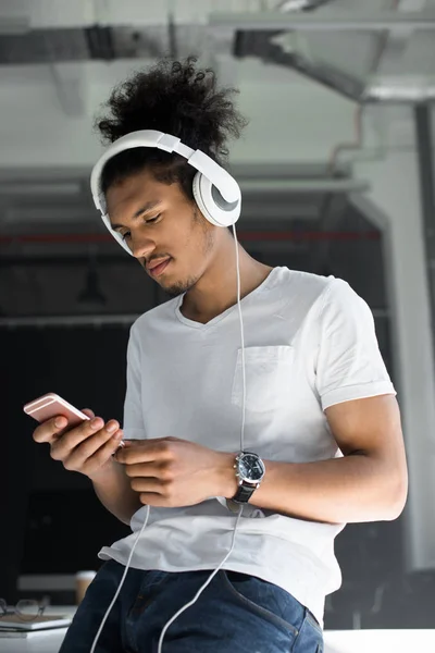 Guapo joven afroamericano hombre en auriculares con teléfono inteligente en la oficina - foto de stock