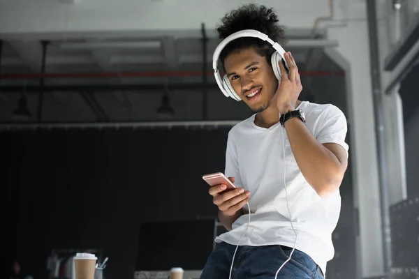 Молодий афроамериканський чоловік в навушниках використовує смартфон і посміхається на камеру в офісі — стокове фото