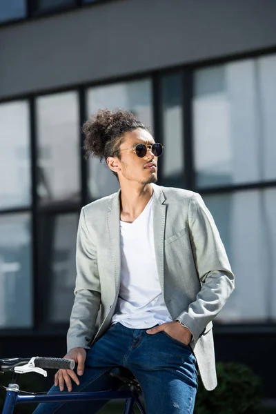 Confiant homme afro-américain élégant dans des lunettes de soleil assis sur le vélo et regardant loin — Photo de stock