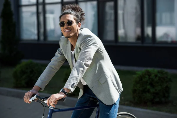 Elegante joven afroamericano hombre en gafas de sol sentado en bicicleta y sonriendo a la cámara - foto de stock