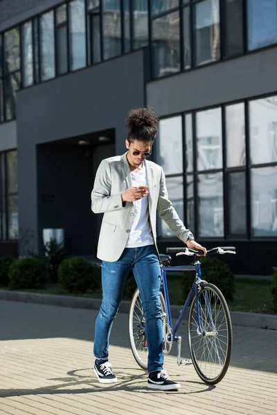 Élégant jeune homme afro-américain en utilisant un smartphone tout en marchant à vélo sur la rue — Photo de stock
