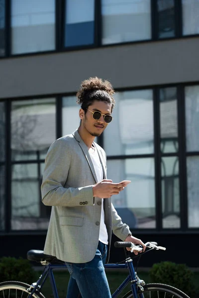 Jovem homem americano africano olhando para a câmera enquanto está de pé com bicicleta e usando smartphone — Stock Photo