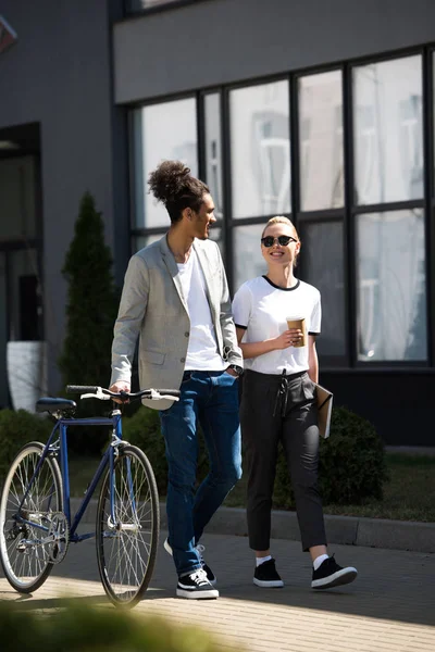 Heureux jeune couple multiethnique regarder l'autre tout en marchant dans la rue avec vélo et café pour aller — Photo de stock