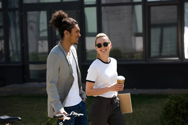 Happy young multiethnic couple talking while walking on street with bicycle and coffee to go — Stock Photo