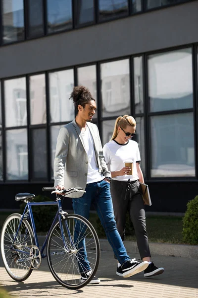 Улыбающаяся молодая многонациональная пара с велосипедом, идущая по улице — стоковое фото