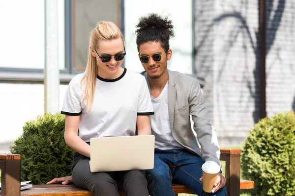 Sorrindo jovem casal multiétnico em óculos de sol usando laptop enquanto sentados juntos no banco — Fotografia de Stock