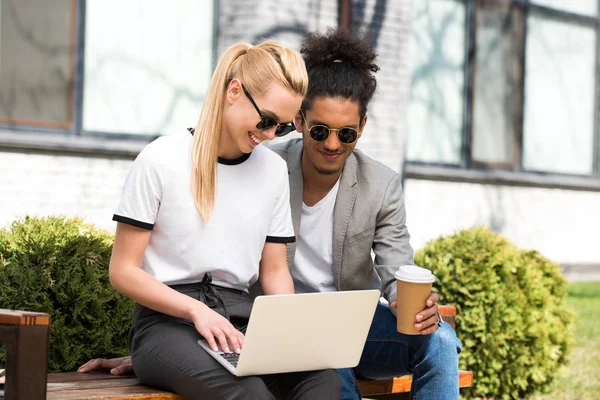 Sourire jeune couple multiethnique en utilisant un ordinateur portable et boire du café pour aller tout en étant assis ensemble sur le banc — Photo de stock