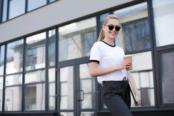 Hermosa chica sonriente sosteniendo café para ir y portátil, mientras que de pie en la calle - foto de stock