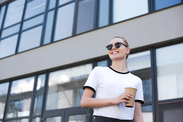Vista basso angolo di bella sorridente giovane donna che tiene il caffè per andare e guardando lontano mentre cammina sulla strada — Foto stock
