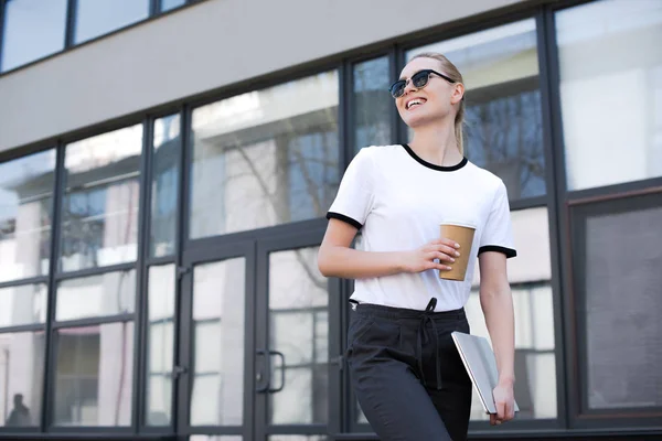 Linda menina loira sorridente com café para ir e laptop andando e olhando para longe na rua — Fotografia de Stock