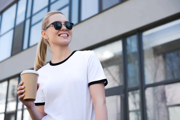 Vista basso angolo di felice giovane donna in occhiali da sole che tiene il caffè per andare e guardando lontano sulla strada — Foto stock