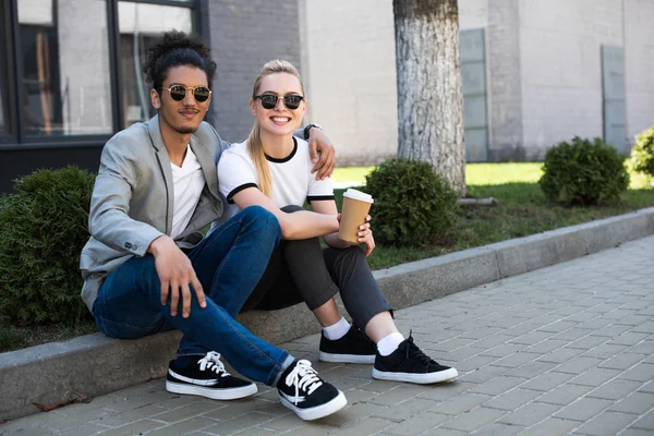 Feliz joven pareja multiétnica sonriendo a la cámara mientras está sentado en la calle con café para ir - foto de stock