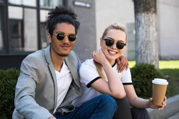 Joven pareja multiétnica en gafas de sol sonriendo a la cámara mientras toma café para salir a la calle - foto de stock