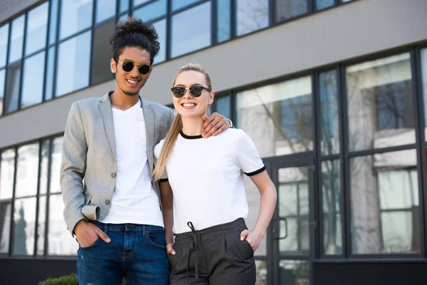 Joven pareja multiétnica en gafas de sol de pie juntos y sonriendo a la cámara en la calle - foto de stock