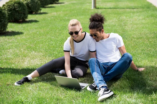 Happy young multiethnic couple sitting on grass and using laptop — Stock Photo