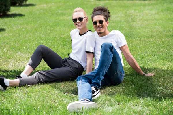 Glückliches junges multiethnisches Paar mit Sonnenbrille, das zusammen auf dem Gras sitzt und in die Kamera lächelt — Stockfoto