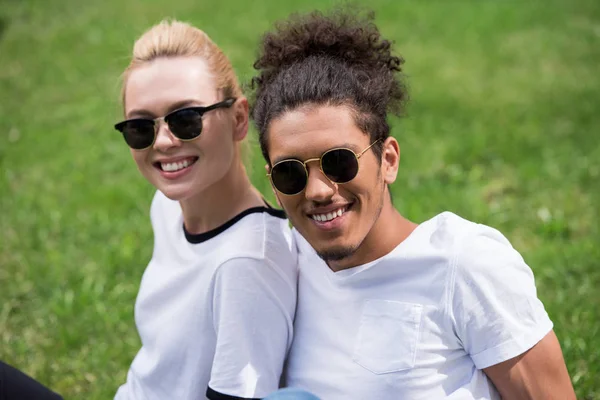 Retrato de belo jovem casal multiétnico em óculos de sol sentados juntos na grama e sorrindo para a câmera — Fotografia de Stock