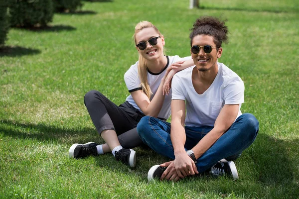 Красивая молодая многонациональная пара в солнечных очках, сидящая вместе на траве и улыбающаяся в камеру — стоковое фото