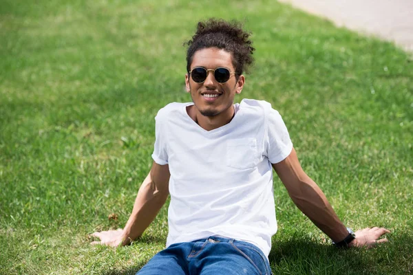 Handsome young african american man in sunglasses sitting on grass and smiling at camera — Stock Photo