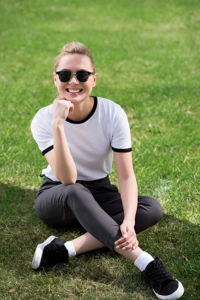 Красива блондинка в сонцезахисних окулярах сидить на траві і посміхається на камеру — стокове фото