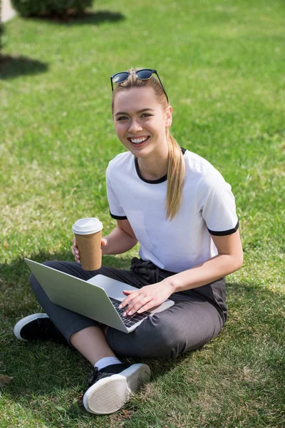 Красива блондинка з кавою, щоб посміхатися на камеру, сидячи на траві і використовуючи ноутбук — стокове фото