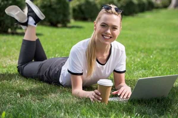 Beautiful blonde girl smiling at camera while lying on grass and using laptop — Stock Photo