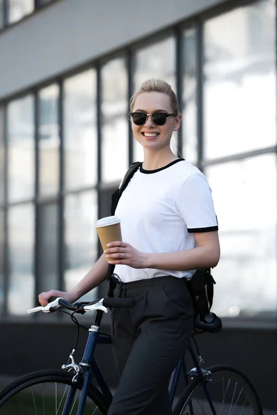 Красивая улыбающаяся девушка в солнечных очках держит бумажную чашку и стоит с велосипедом на улице — стоковое фото