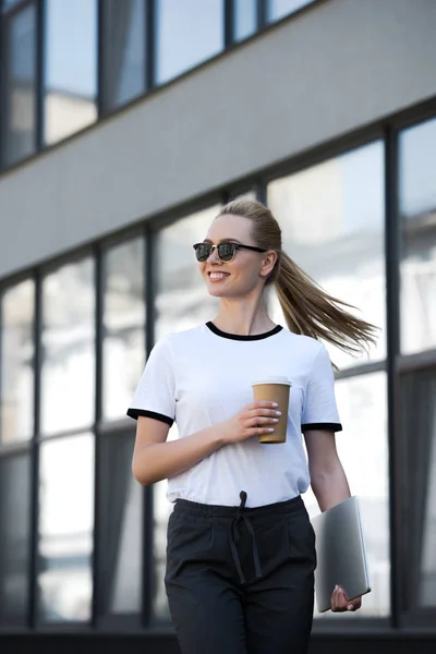 Chica rubia sonriente con ordenador portátil y taza de café desechable mirando hacia el exterior edificio de oficinas - foto de stock