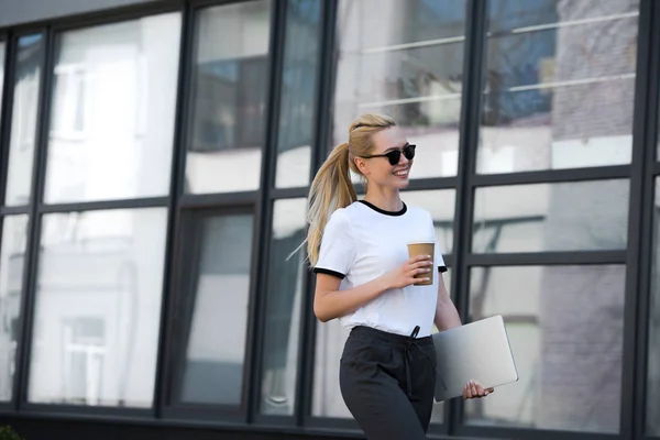 Красива усміхнена дівчина в сонцезахисних окулярах тримає паперовий стаканчик і ноутбук під час виходу за межі офісної будівлі — стокове фото