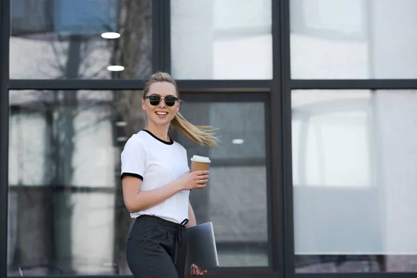 Belle jeune femme en lunettes de soleil souriant à la caméra tout en tenant tasse en papier et ordinateur portable à l'extérieur de l'immeuble de bureaux — Photo de stock