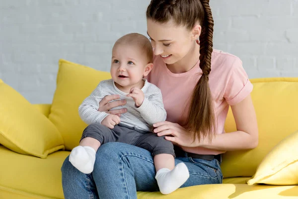 Madre joven sentada en un sofá con un bebé pequeño - foto de stock
