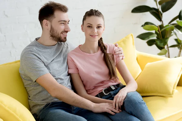 Jovem sorridente homem e mulher abraçando e sentado no sofá — Fotografia de Stock