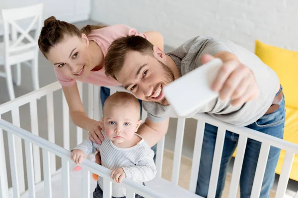 Smiling parents taking selfie with son in baby crib on smartphone — Stock Photo