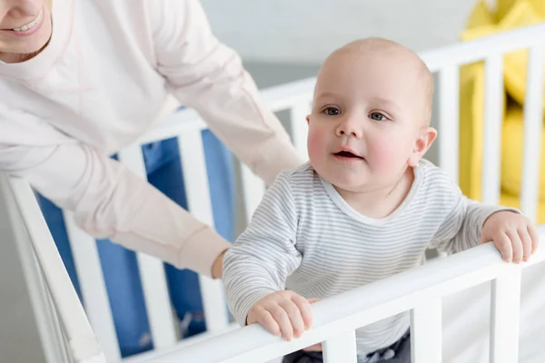 Little son standing in baby crib with mother behind — Stock Photo