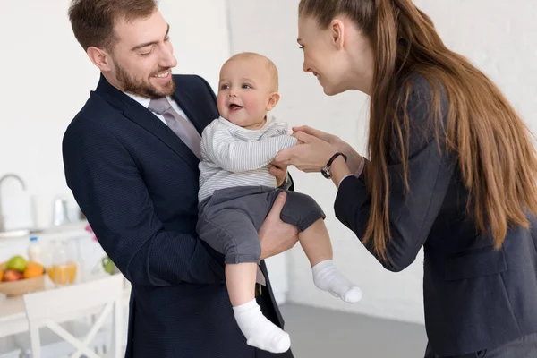 Sonrientes padres de negocios en ropa formal sosteniendo al bebé en casa - foto de stock