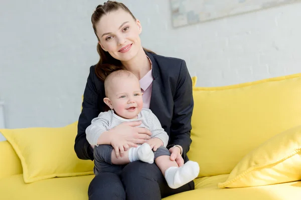 Feliz mujer de negocios en traje con pequeño hijo sentado en el sofá - foto de stock