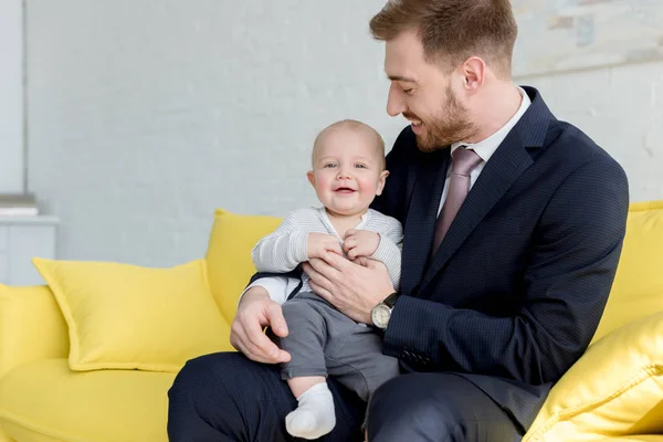 Felice uomo d'affari in giacca e cravatta seduto sul divano con piccolo figlio — Foto stock